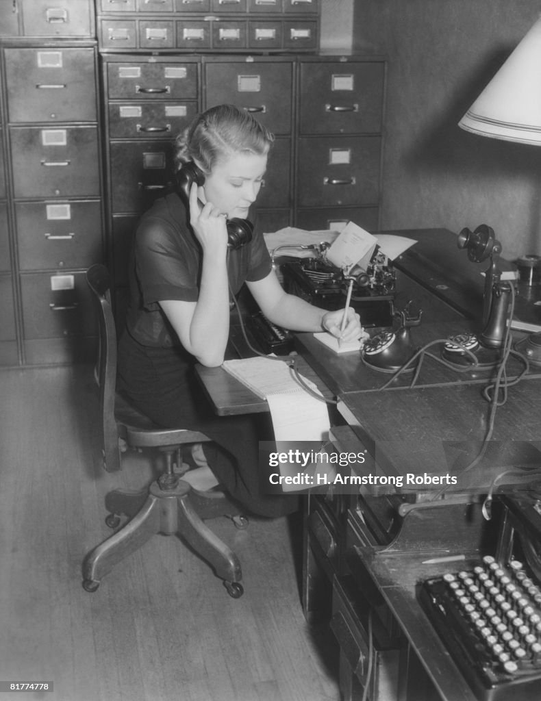 Secretary sitting at desk behind typewriter, on phone taking message.