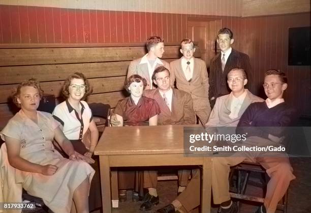 High school students sit at a small table in a gymnasium during a high school dance, with two people in a couple and others smiling or standing...