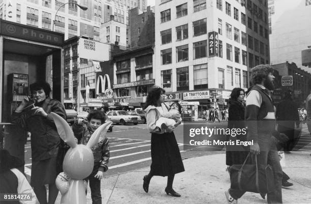 Woman talks on the telephone, cigarette in hand, while a young boy plays with a large, inflatable rabbit and other pedestrians walk past on an...