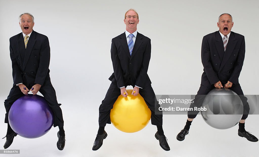 Three mature businessmen bouncing on space hoppers, laughing