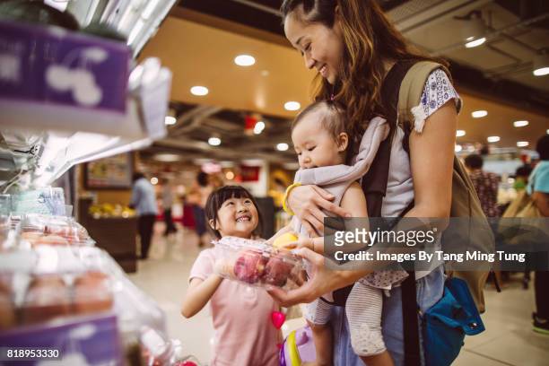 Pretty young mom shopping with her lovely little daughter in the supermarket joyfully while carrying her baby with a baby carrier.