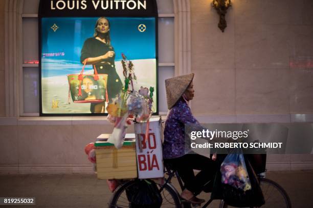 In this photograph taken on July 19 a bicycle vendor offering her merchandise in the streets of Hanoi pedals past a high end merchandise mall in the...