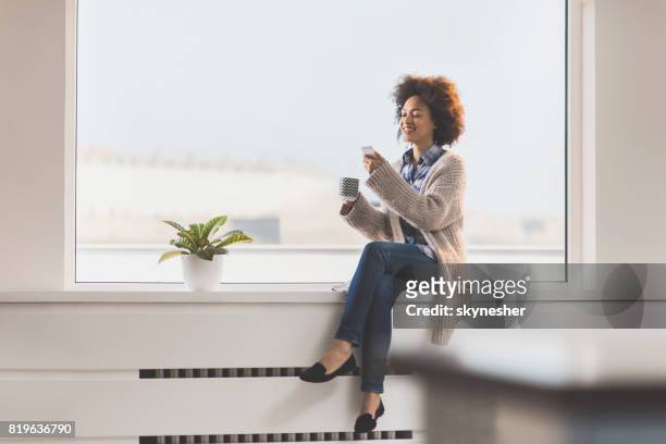 beautiful african american woman using mobile phone while relaxing on a window sill. - windowsill copy space stock pictures, royalty-free photos & images