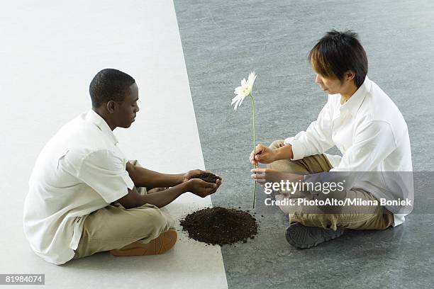 two males sitting face to face on ground, one holding out handful of soil, the other holding out a flower - man holding out flowers stock pictures, royalty-free photos & images