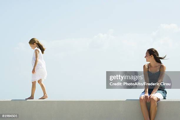 young woman sitting outdoors, watching younger sister walk across ledge - ledge stock pictures, royalty-free photos & images