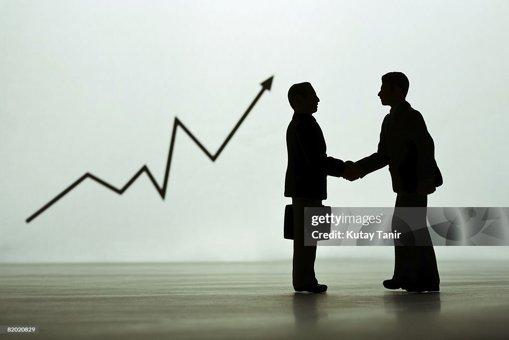 Silhouette of businessmen figurines shaking hands, graph in background. (Focus on foreground)