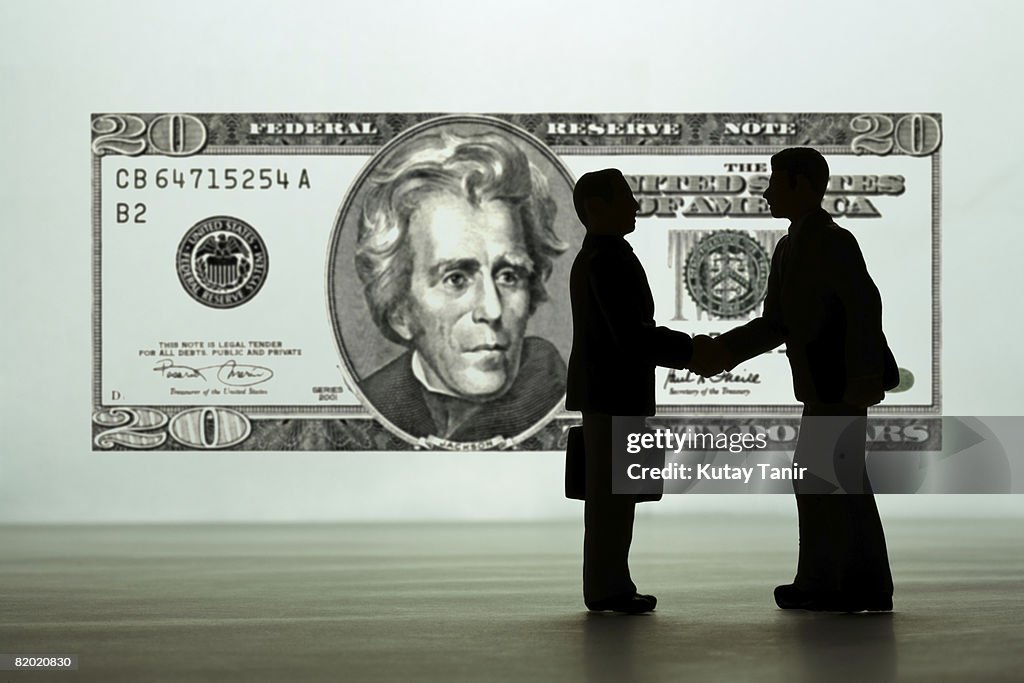 Silhouette of businessmen figurines shaking hands, dollar bill in background. (Focus on foreground)