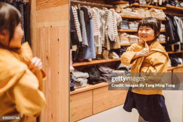 lovely little girl trying out a jacket in front of a mirror joyfully in a department store - girl changing room shop stock-fotos und bilder