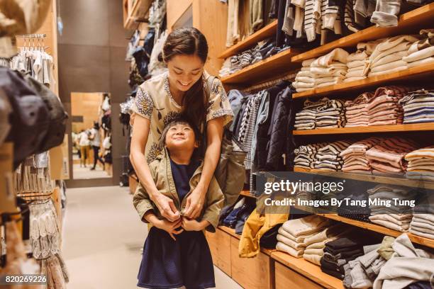 pretty young mom trying out a jacket on her lovely little daughter in a department store joyfully. - shopping candid stock pictures, royalty-free photos & images