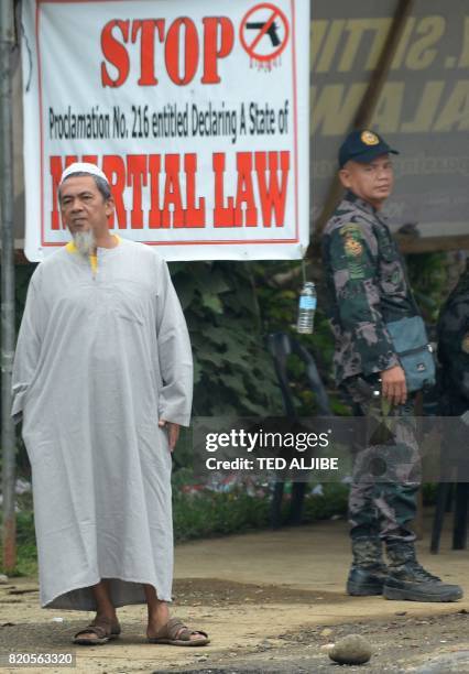 Resident stands next to military troops guarding a checkpoint with a tarpaulin notifying motorists and residents of the implementation of martial...