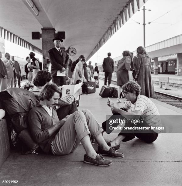 Three young adults sitting on the ground on a platform of West Station in Vienna, Elder travellers in the background, Photograph, 1975 [Drei junge...