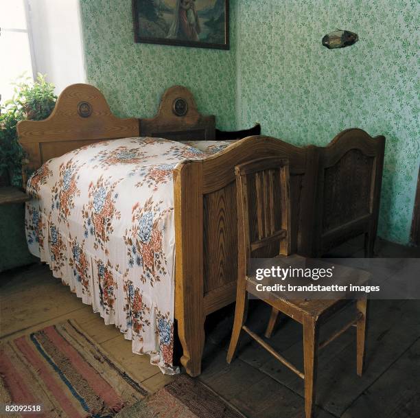 Wooden peasant full-size bed with coverlet with floral design and a wooden chair standing in front of the bed, Bedroom in a homestead in Streith in...