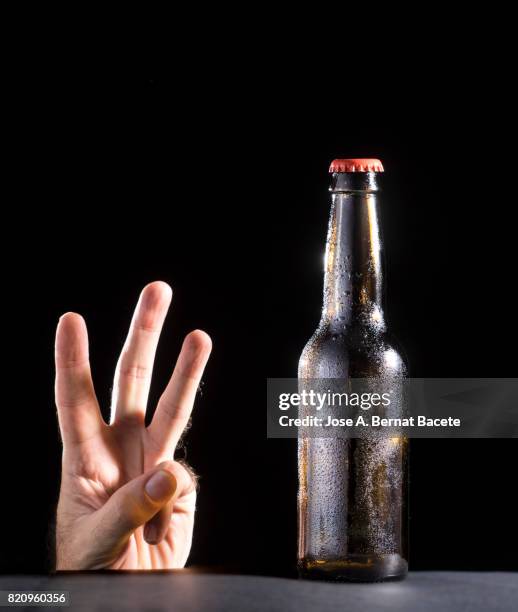 bottle of beer with the glass esmerilado with drops of water and the hand of a man with a hand gesture of three fingers on a black bottom - beer mat stockfoto's en -beelden