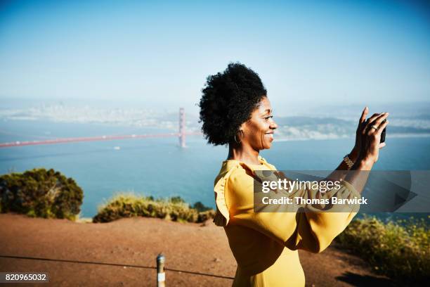 smiling woman taking photo with smartphone while standing at vista above san francisco - tour of california stock-fotos und bilder