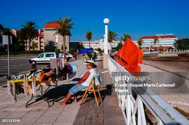 Woman sitting at a souvenirs street stall on beach promenade and Hotel & Talaso Louxo La Toja at back.