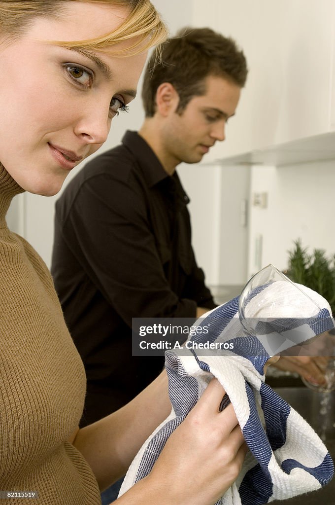 Side profile of a young woman rubbing a wine glass with a dish towel and a young man standing beside her
