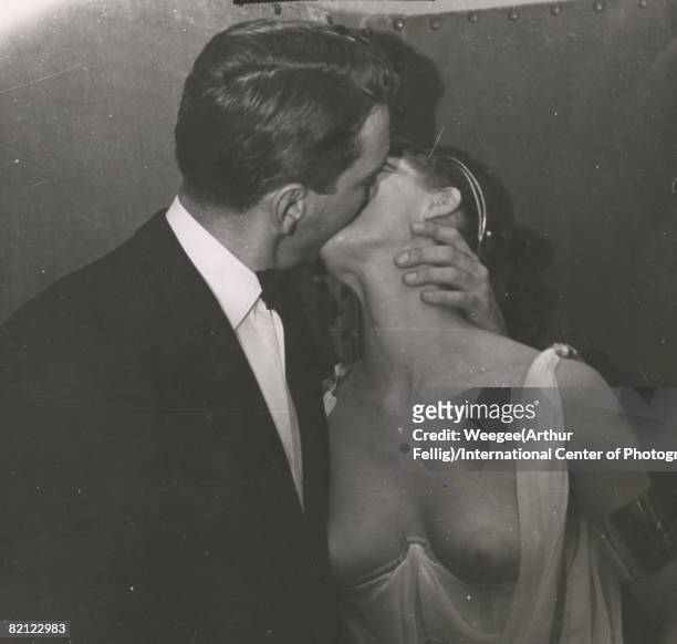 Portrait of a man and woman as they kiss in a corner, 1950s. The man in dressed in a suit and tie, the woman in a chiffon robe that leaves her...