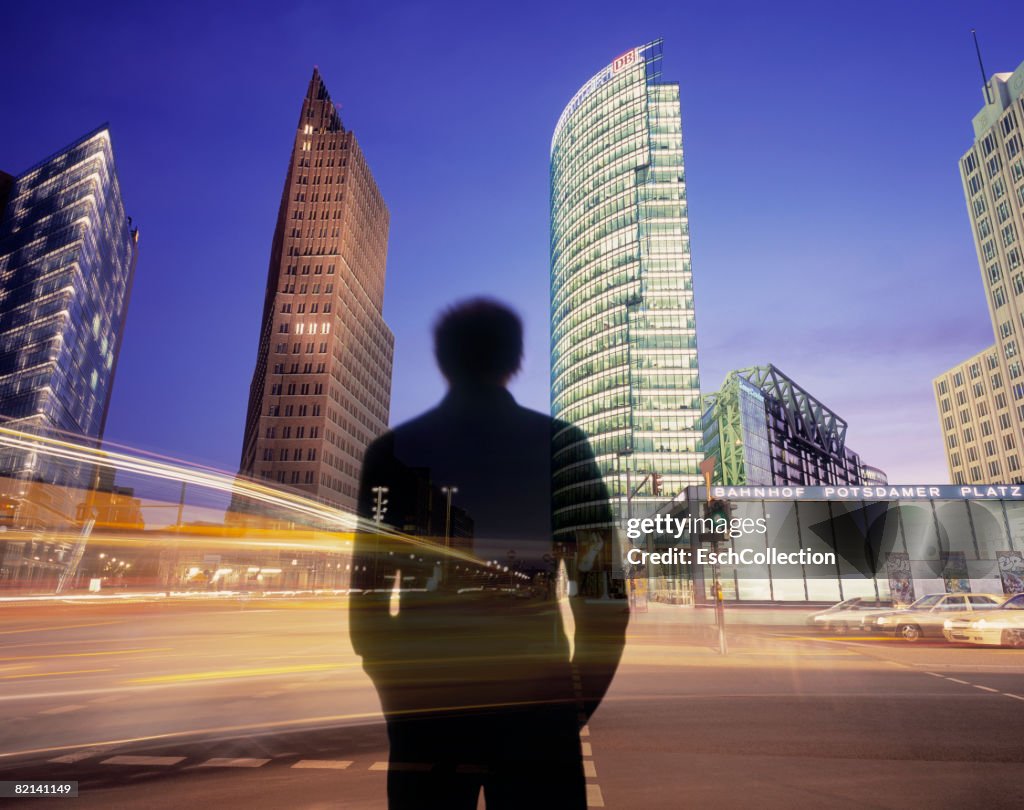 Man waiting to cross busy road with illuminated offices.