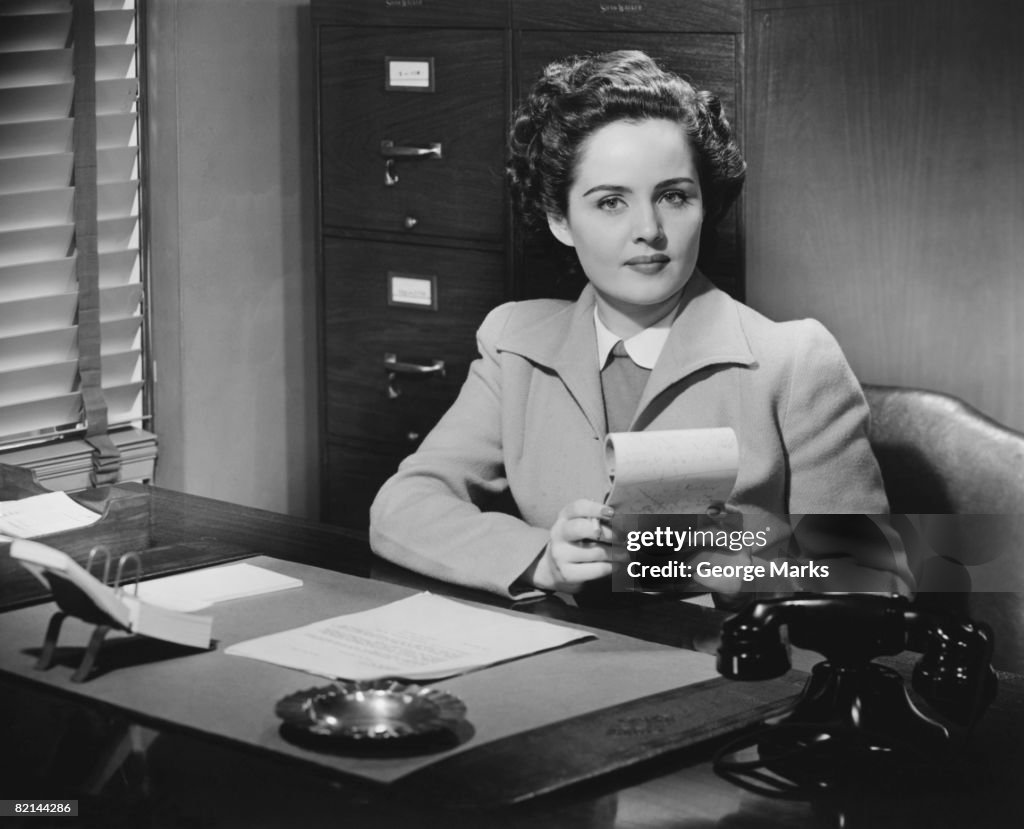 Businesswoman sitting at desk, holding notepad, (B&W), portrait