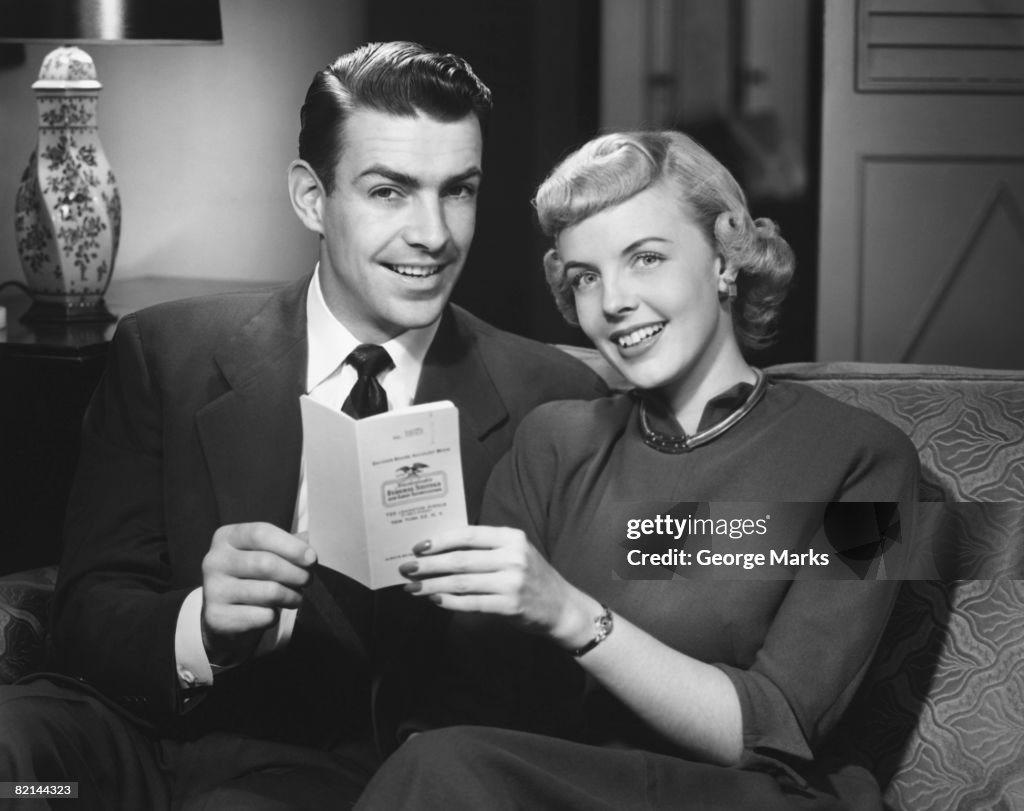 Couple sitting on sofa in living room, holding bank book, (B&W), portrait