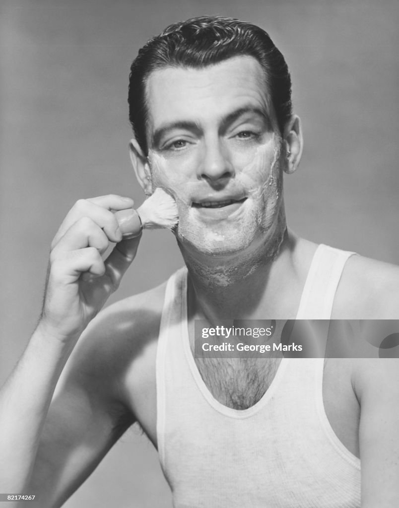 Man applying shave foam on face in studio, (B&W), portrait