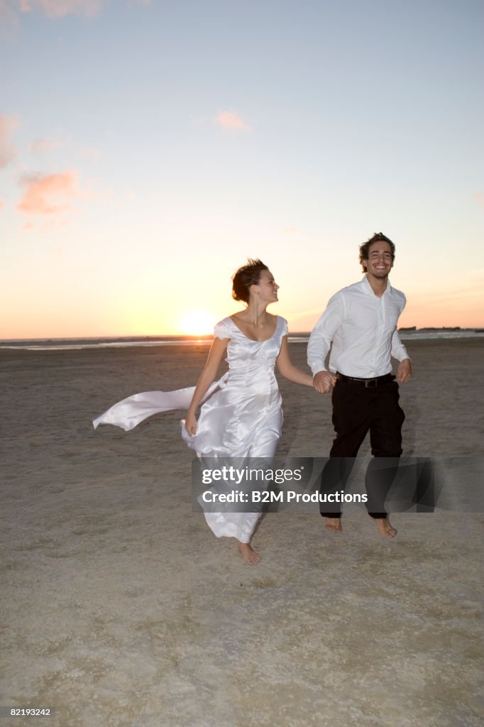 Bride and groom running on beach holding ha