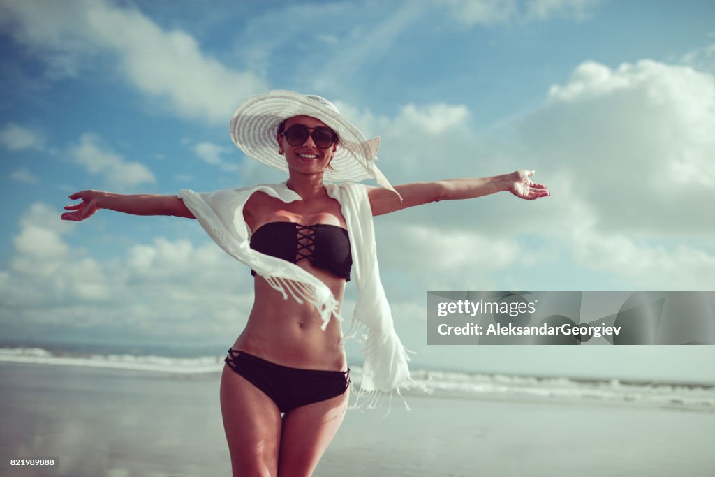 Lächelnde junge Frau mit Hut und Sonnenbrille posiert mit erhobenen Händen am Ocean Beach in Bali