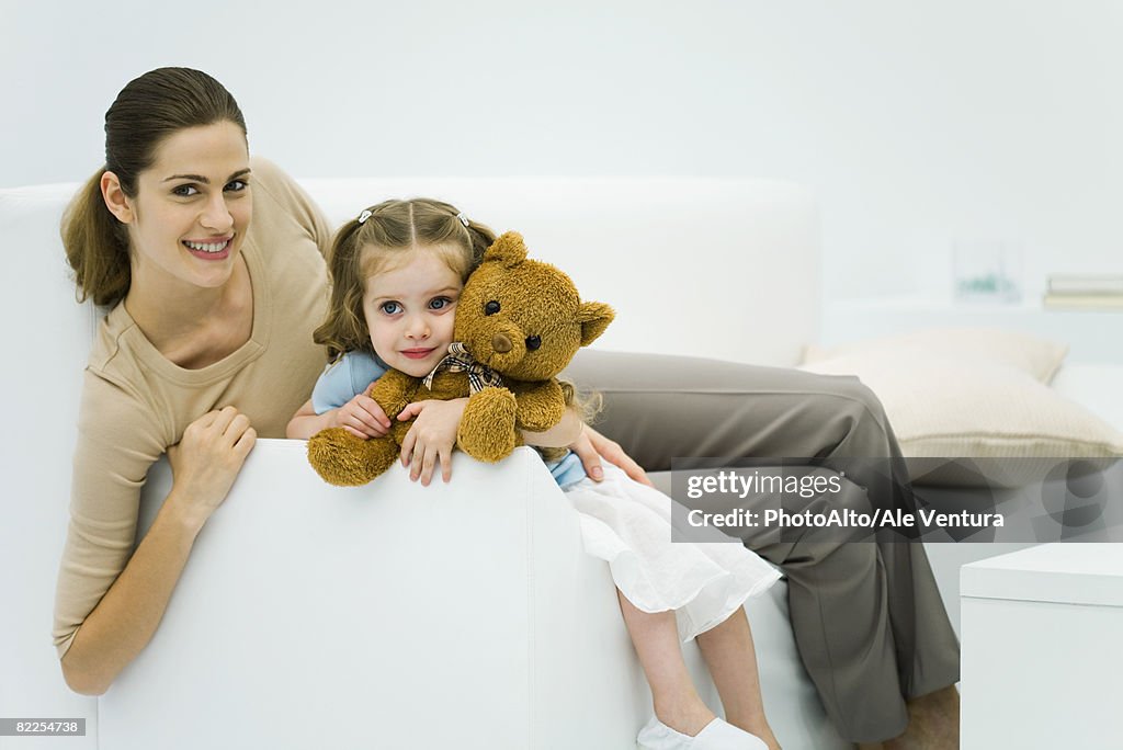 Mother and daughter sitting on couch, woman smiling at camera, girl holding teddy bear