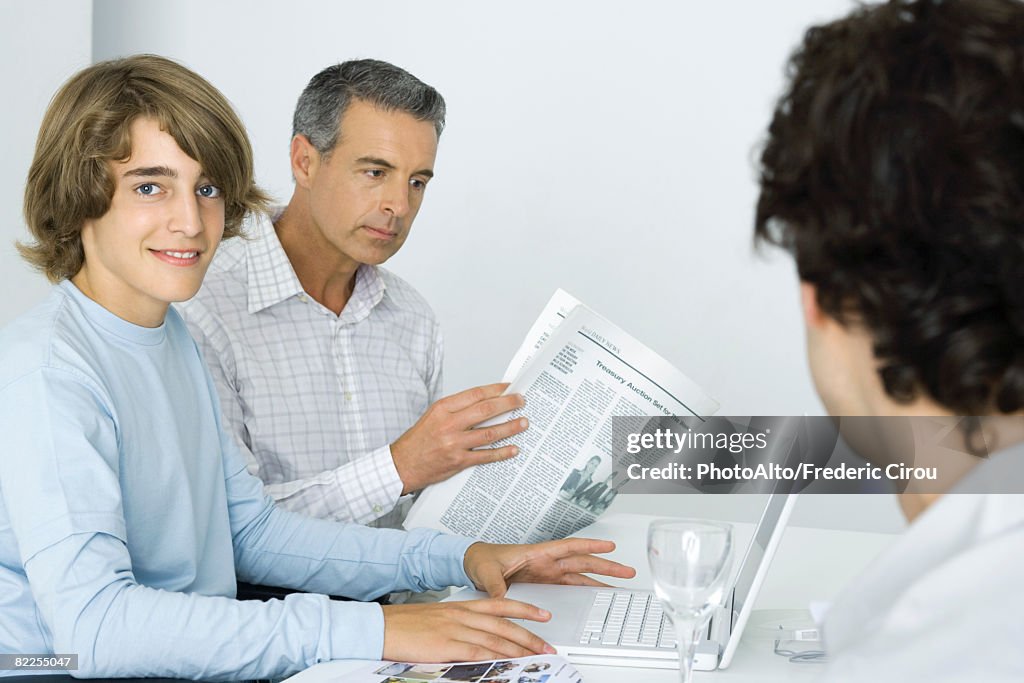 Teen boy at table with father, using laptop computer, man reading newspaper