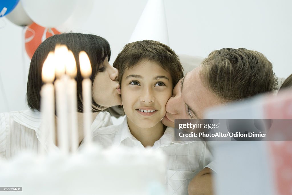 Family behind birthday cake with lit candles, parents kissing boy on cheeks