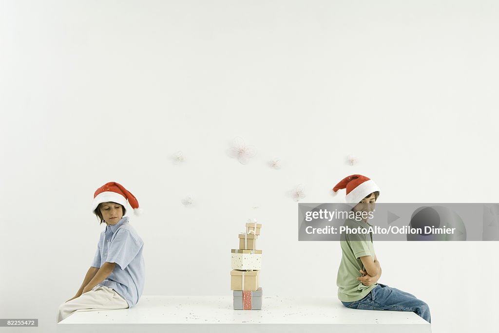 Two brothers sitting back to back, Christmas gifts stacked between them, both wearing Santa hats