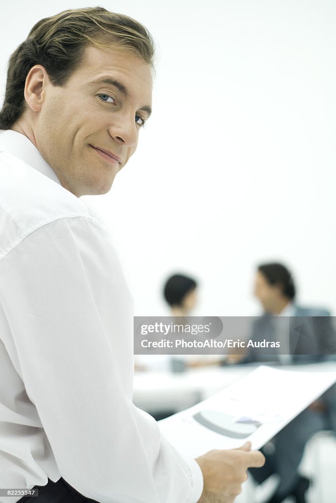 Businessman smiling over shoulder at camera, holding document, colleagues in background