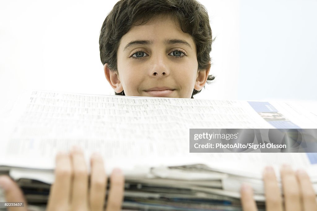 Boy holding tall stack of newspapers, smiling at camera