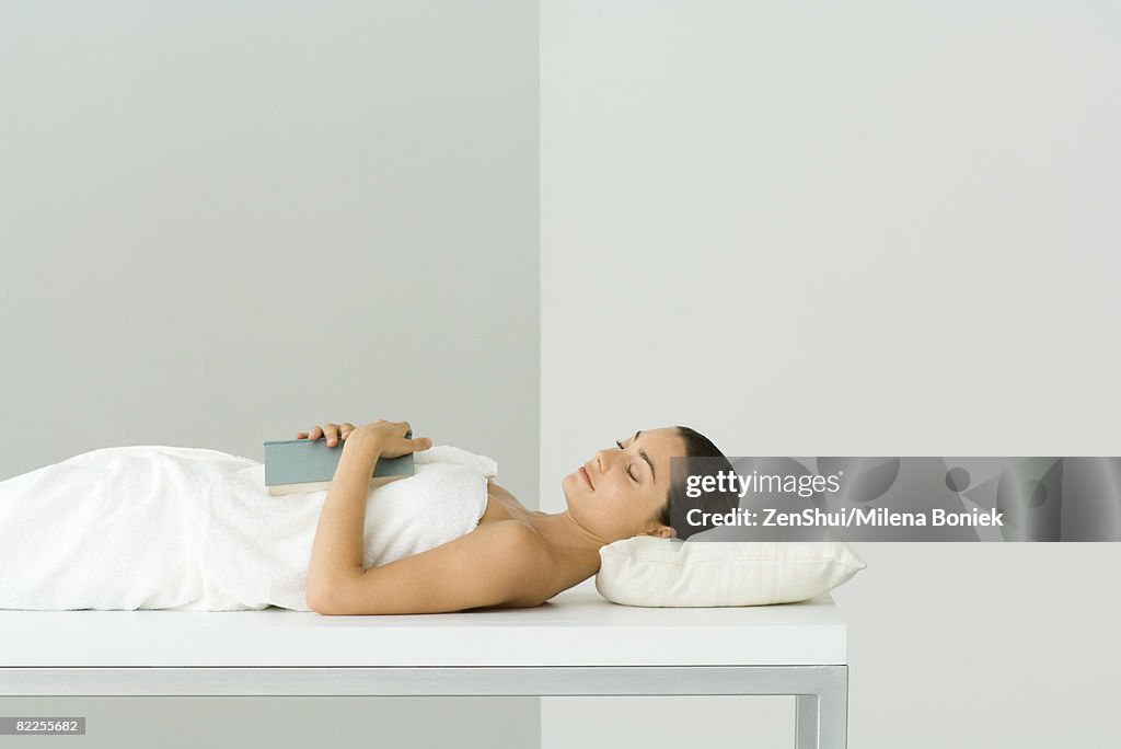 Woman lying on massage table, holding book on chest, eyes closed