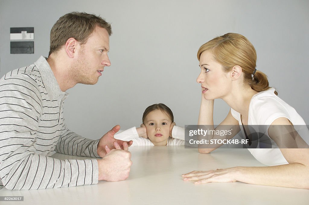 Couple arguing at table, girl in background covering ears with hands