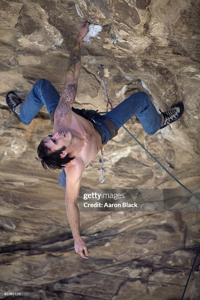 Man wearing blue jeans, without shirt, with tattoos hangs upside down from one arm while climbing an overhang.