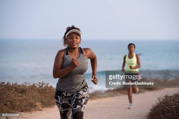 joggen vrouwen uitgevoerd. beginner mollige vrouw en vrouwelijke pro loper tijdens een outdoor workout op de kustlijn. - fat loss training stockfoto's en -beelden