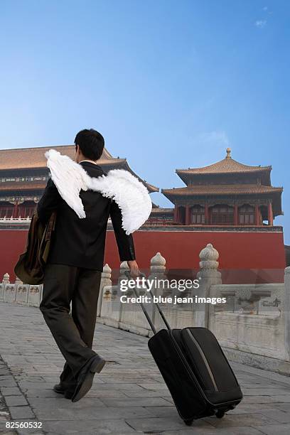 a man in business attire with wings, pulls a piece of luggage, chinese architectural elements behind. - anjo da guarda imagens e fotografias de stock