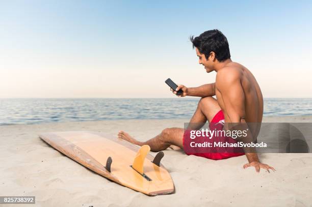 hispanic surfer checking his smart phone - board shorts stock pictures, royalty-free photos & images