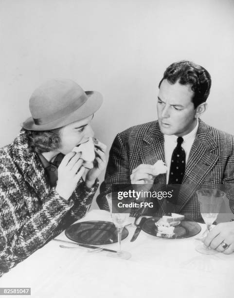 Man expresses disapproval at his friend's table manners as she sinks her teeth greedily into a sandwich, circa 1940.