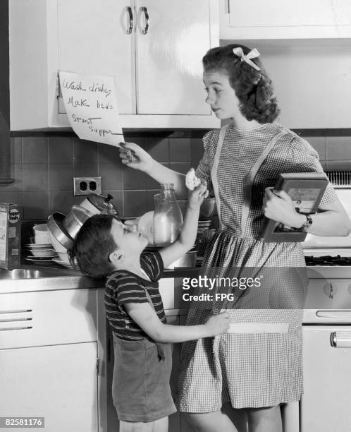 Circa 1950, A teenage girl reads a note with the household chores listed on it, while her younger brother holds up a bandaged finger for attention.