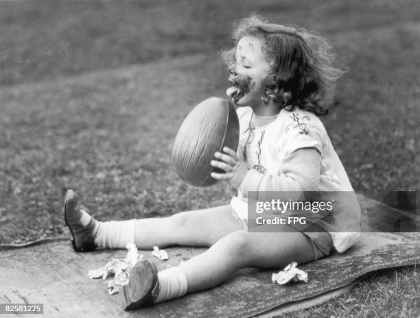 Circa 1930, A little girl makes a pig of herself by licking a large chocolate Easter egg.
