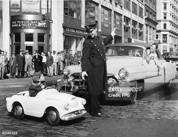 In a staged promotional shot a crowd watches as a policeman stops traffic for six year old Brian M. Dunne as the boy pilots a new fiberglass smallfry...