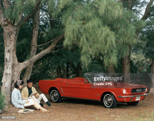 Promotional shot of a red 1964 Ford Mustang convertible parked in a forest clearing with a couple sitting by a pinetree, 1964. This first series of...