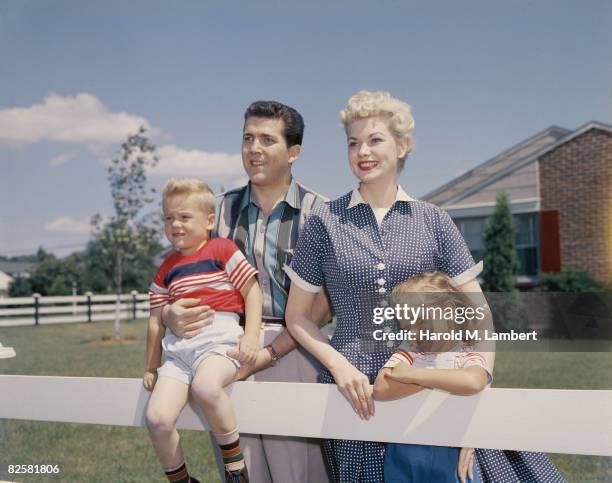 Cheerful young family leaning on the white fence in their garden on a windy day, 1957.