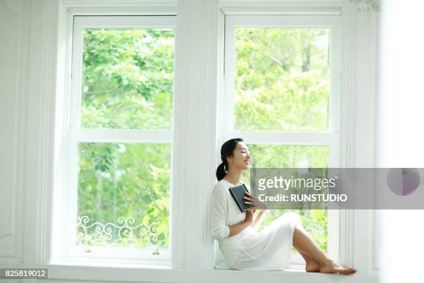 woman sitting in a window and holding a book closing her eyes - closing book stock pictures, royalty-free photos & images