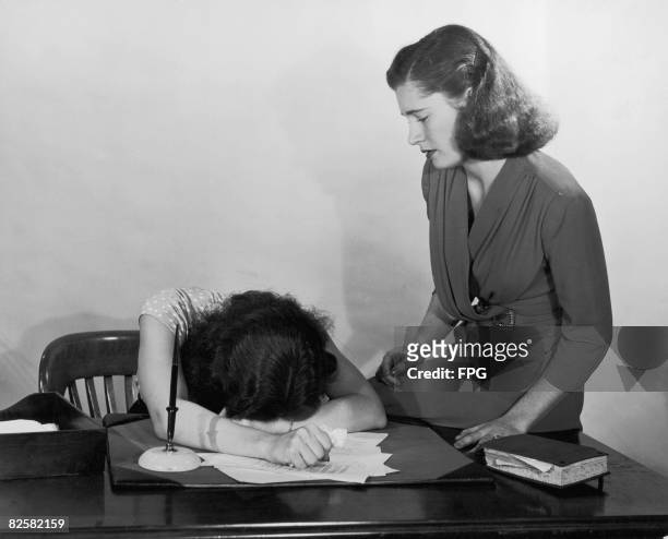 Woman moves to comfort a co-worker who is slumped over her desk in despair, circa 1940.