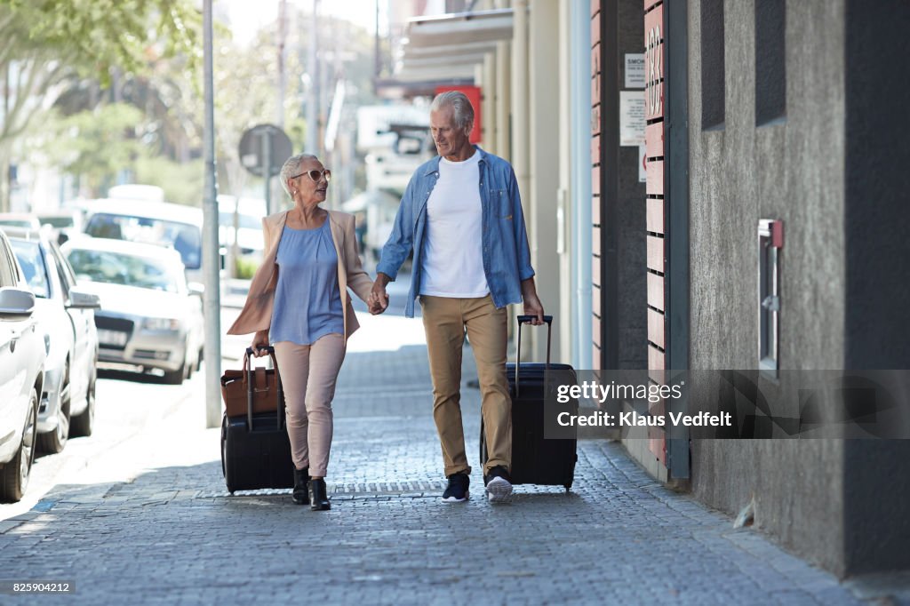 Senior couple walking and talking with rolling suitcases on city street