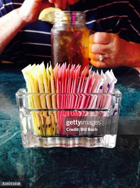 closeup of a glass dish of various artificial sweeteners with woman drinking ice tea in background - artificial sweetener imagens e fotografias de stock