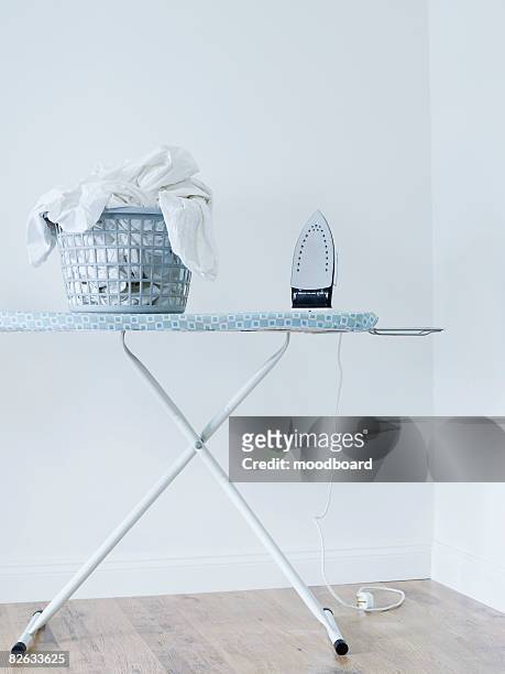 iron and laundry basket on ironing board against white wall - bügelbrett stock-fotos und bilder